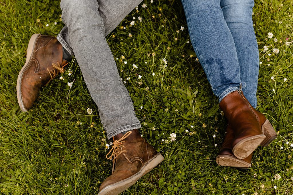 What to wear for my engagement session couple sitting in the grass side by side wearing blue and grey jeans and brown shoes