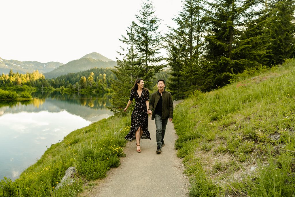 What to wear for my engagement session couple running toward the camera hand in hand, smiling, woman wearing black floral dress, man in jeans and a shirt with bomber jacket