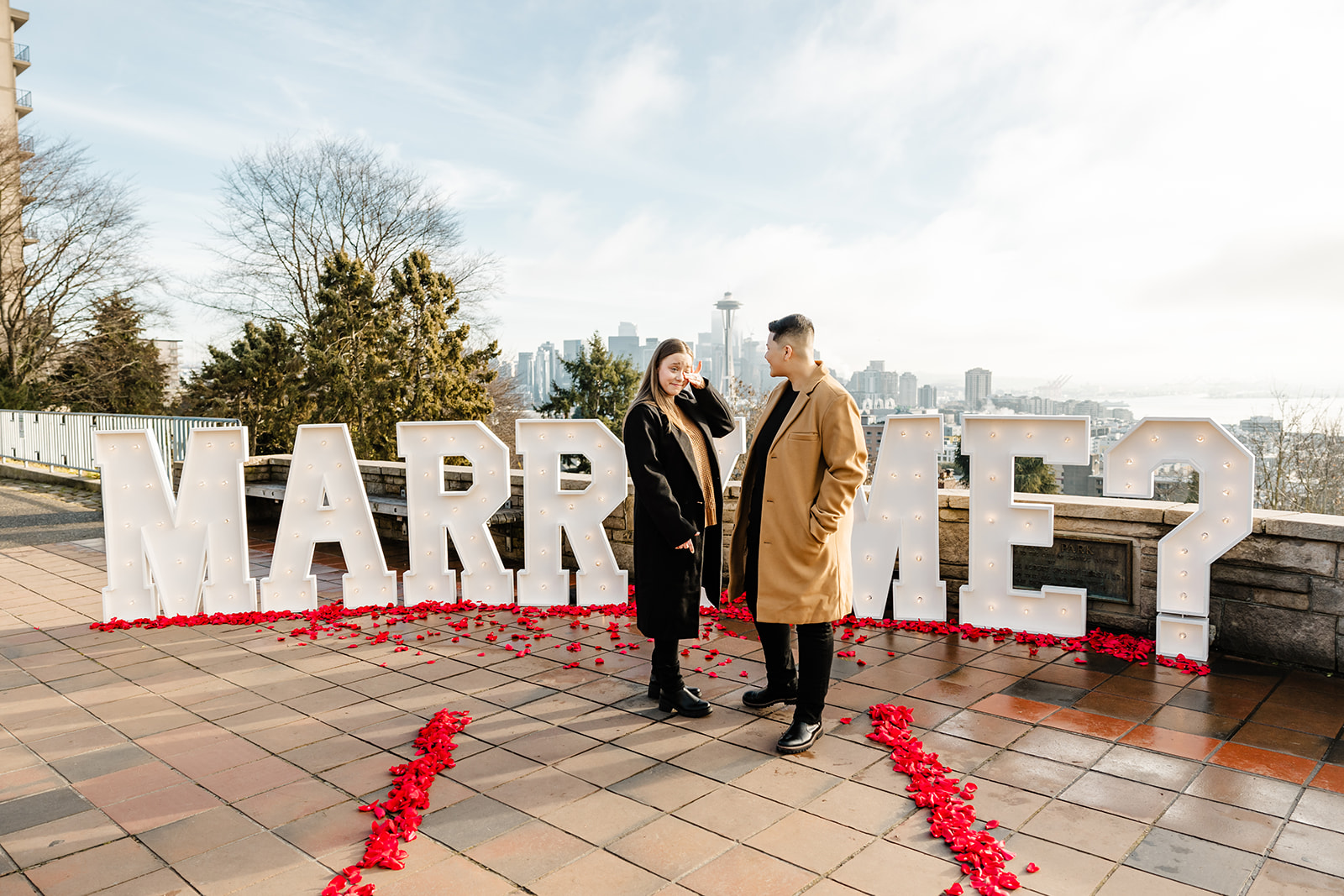 couple standing in front of marry me sign and the seattle skyline at kerry park after proposal, rose petals leading up to engagement spot