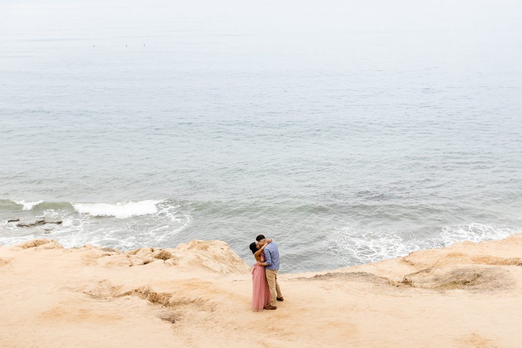 sunset cliffs engagement photographer couple kissing on the cliffside with ocean in the background, woman wearing prink tulle maxi dress, man wearing blue shirt and khakis, What to wear for my engagement session
