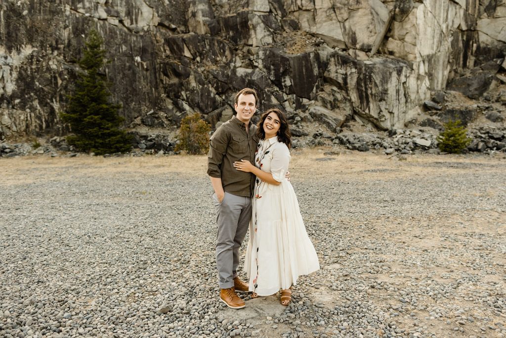 couple standing side by side smiling at the camera, woman wearing a white dress with embroidery and man wearing olive green button up shirt with grey pants