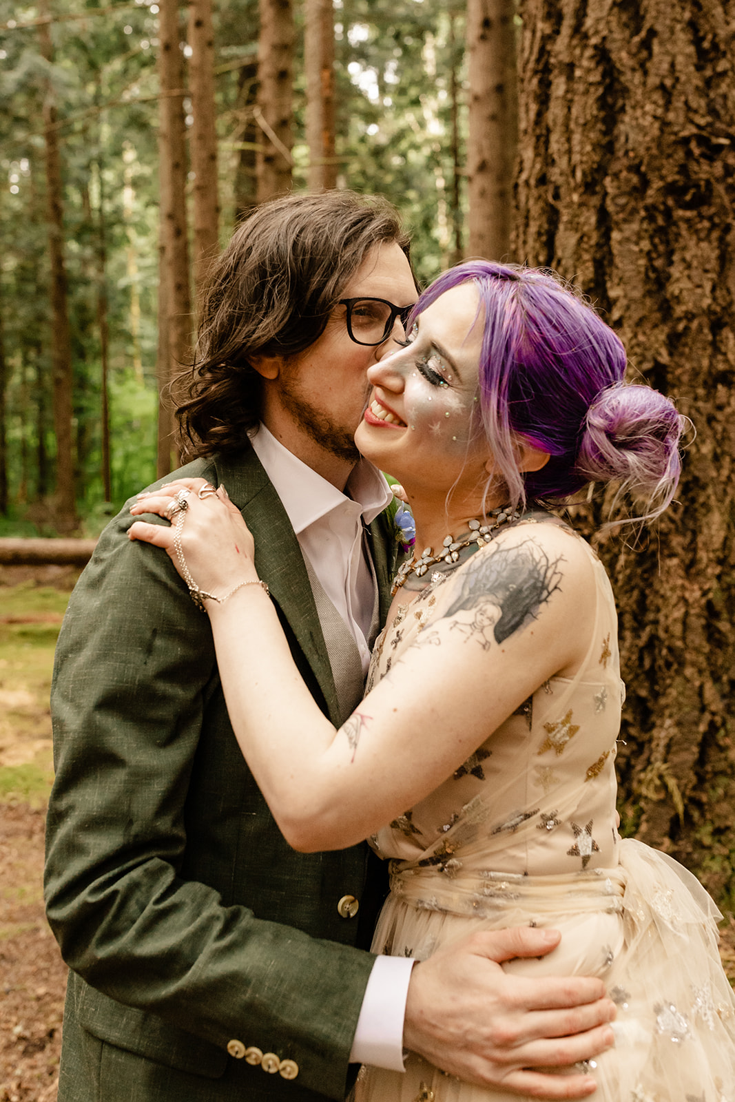 Groom kisses bride on the cheek in the forest.