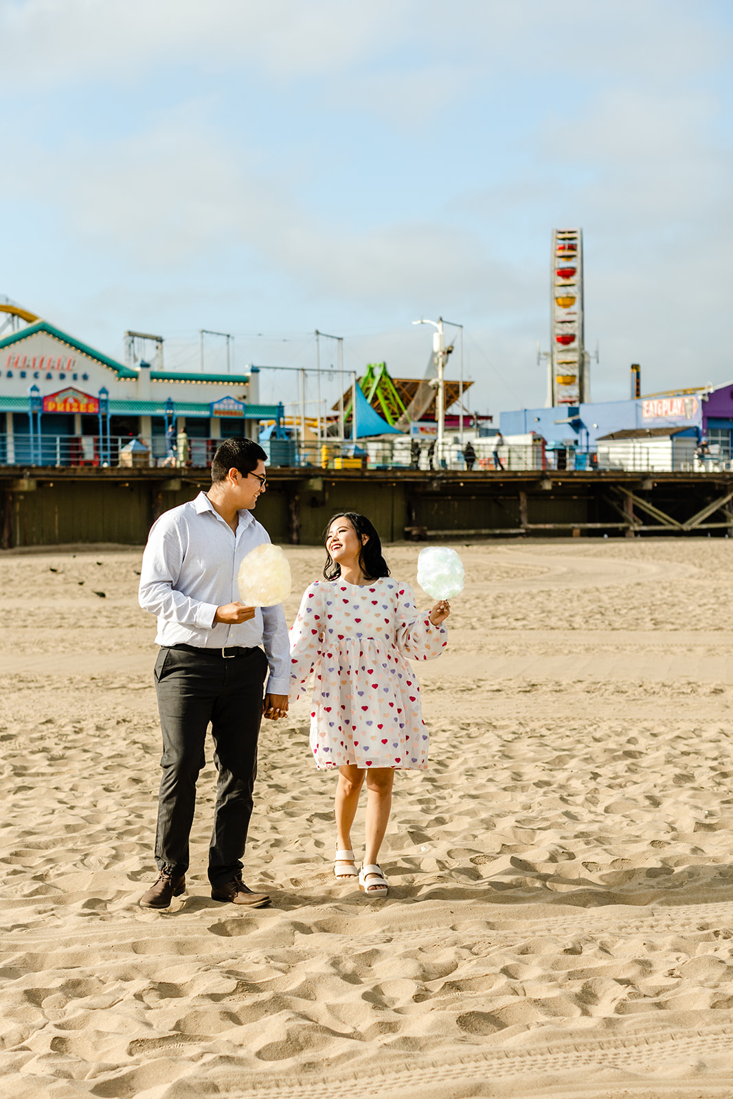 Couple holds hands and hold cotton candy, walking toward camera with Santa Monica Pier in the background.