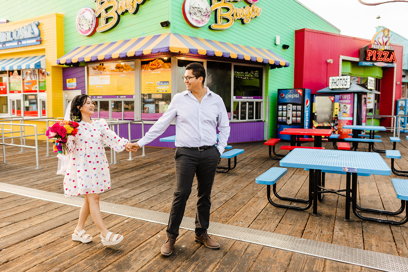 Couple holds hands and walks across frame from left to right on the pier during Santa Monica beach wedding. 