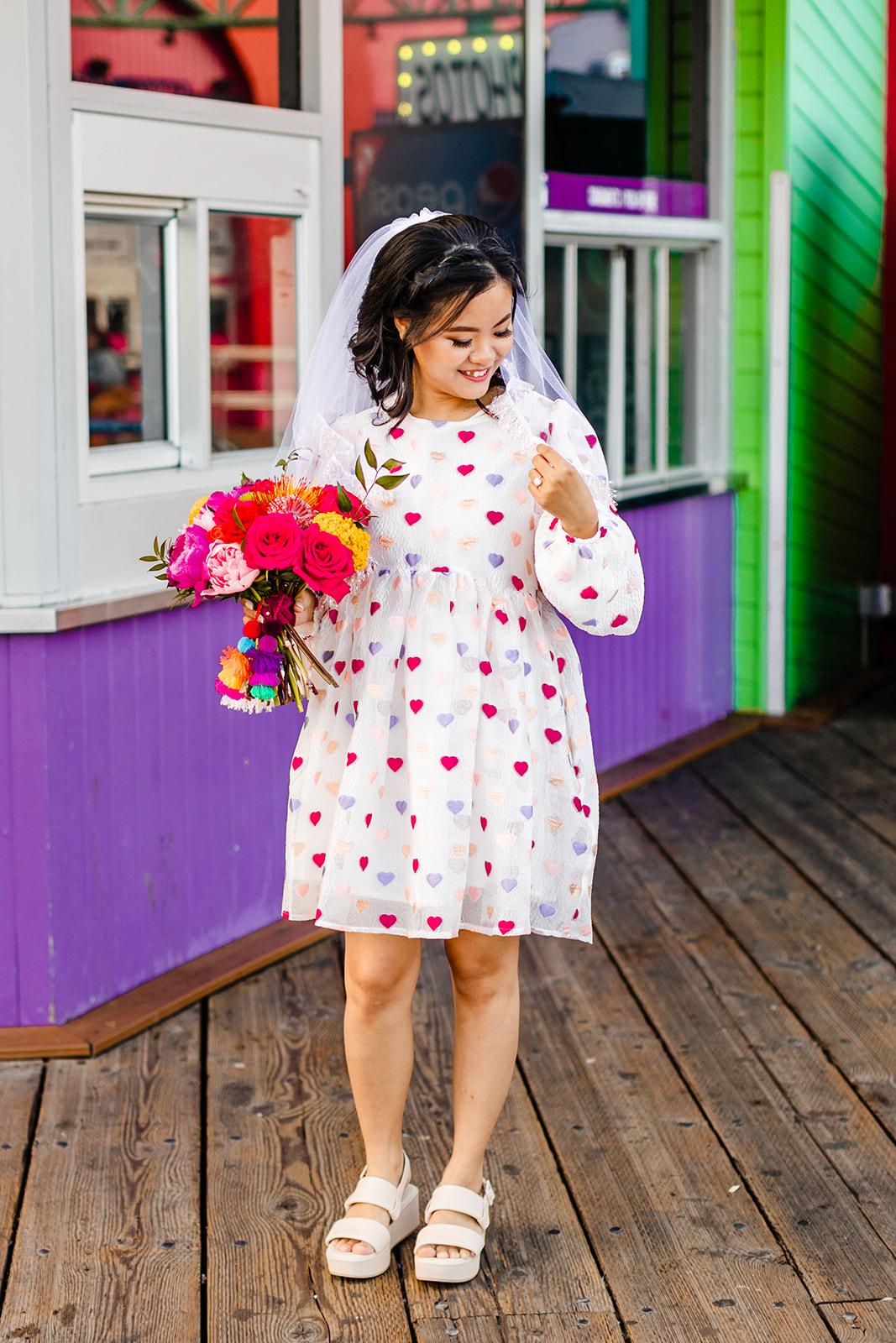 Vertical image of bride holding her bouquet, smiling, touching her veil. She wears a short dress with pink, purple, and red hearts. 