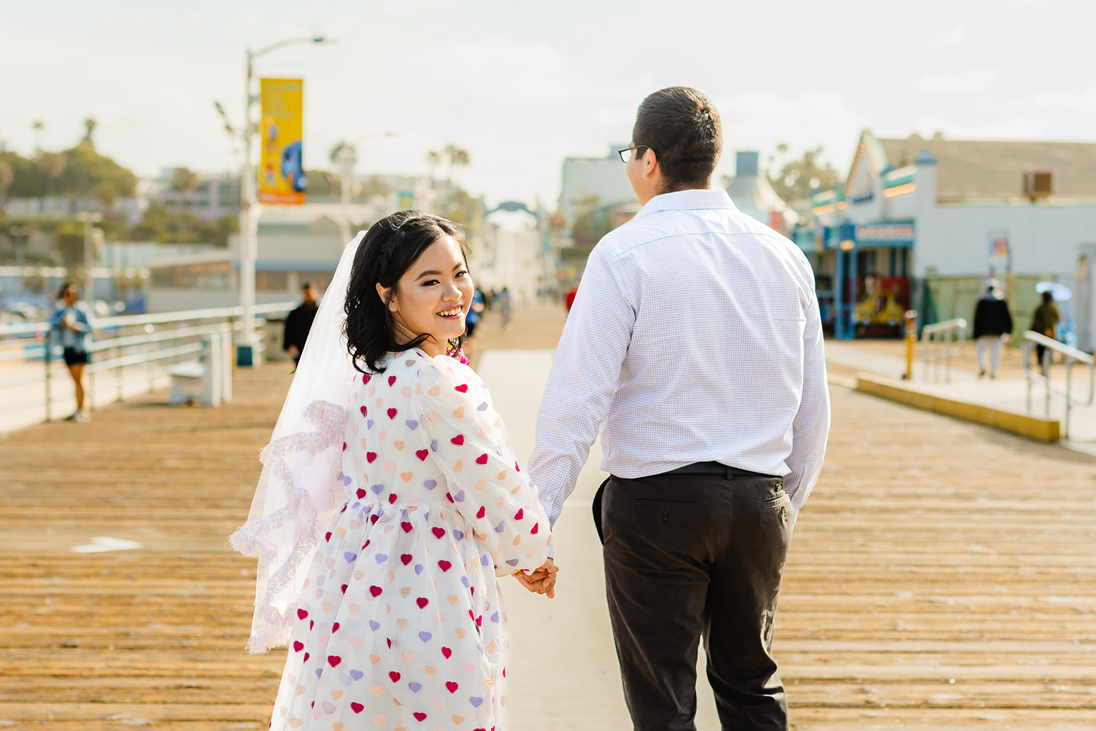 Bride looks over her shoulder at the camera, holding hands with groom. Couple walks down the pier. 