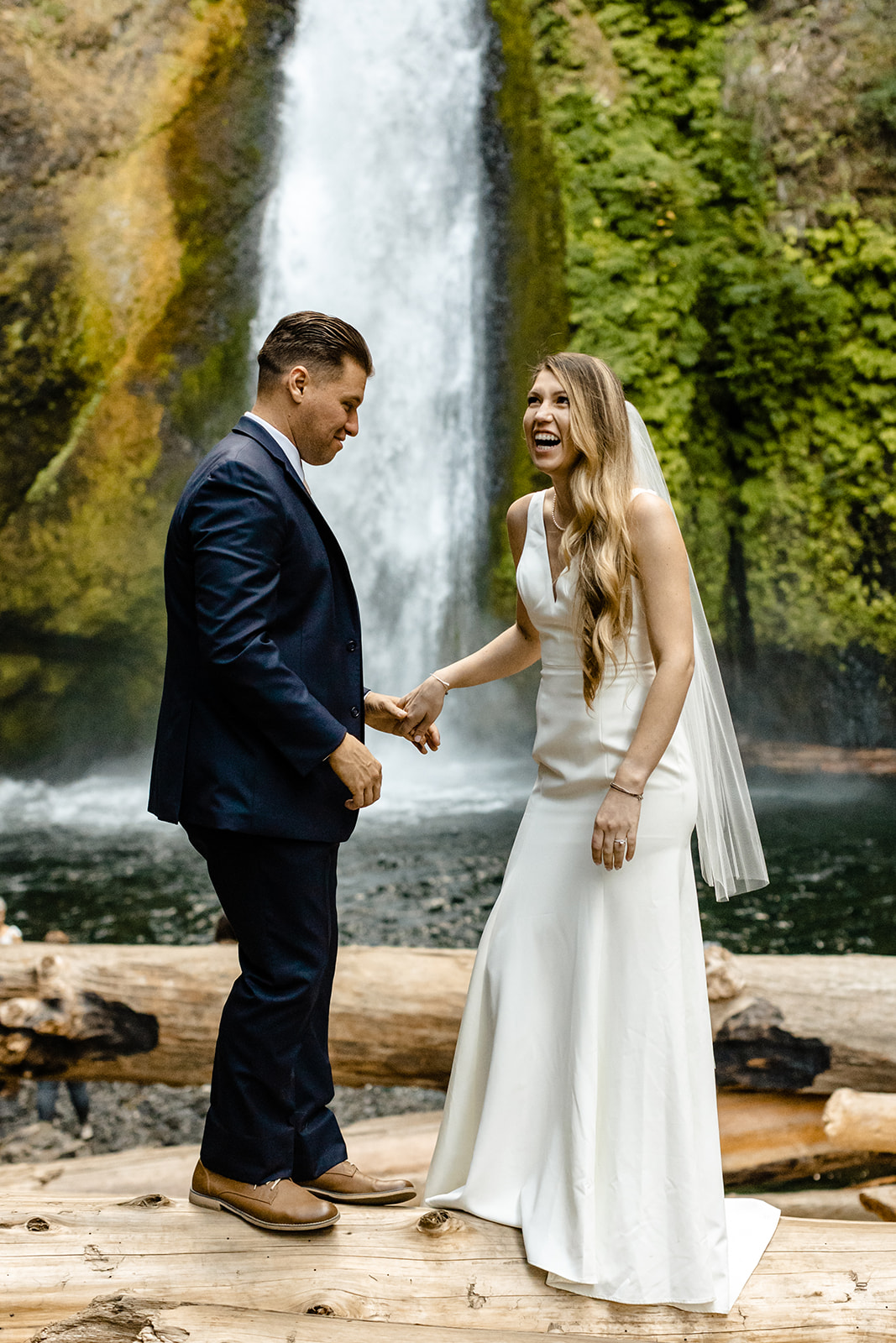 couple balances on fallen logs in front of a waterfall, holding hands.
