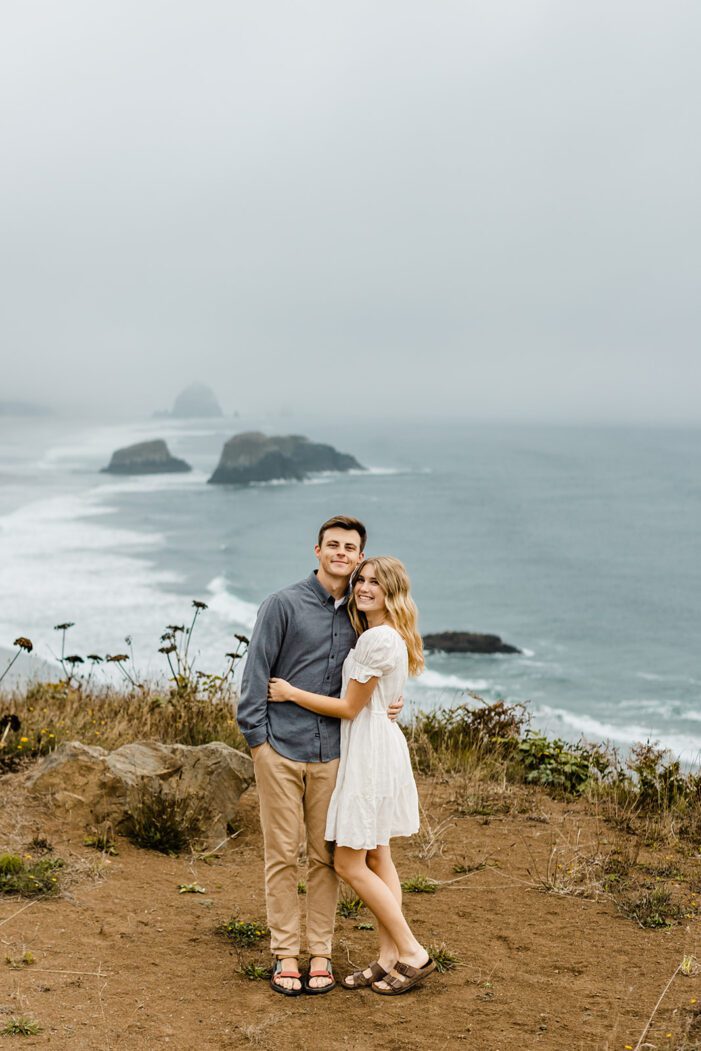 boy and girl stand on cliffside during cannon beach engagement
