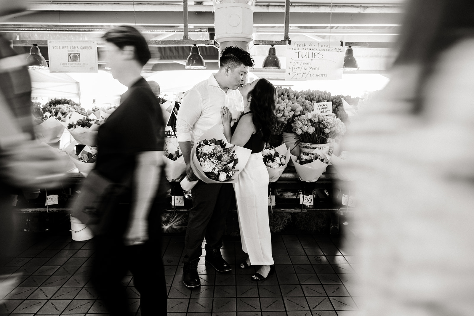 people walk in front of bride and groom posing in front of flower stand at pike place market