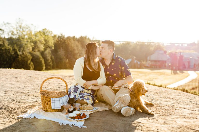 couple sits on picnic blanket and kiss with their dog beside them