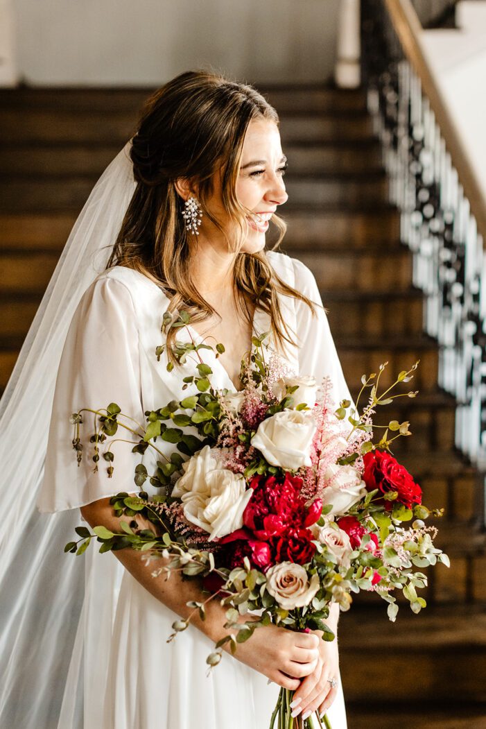bride stands in front stairs holding pink and red bouquet