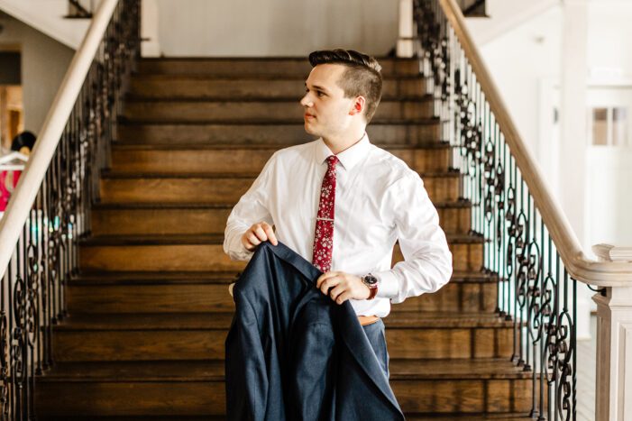 groom stands in front of stairs putting on jacket