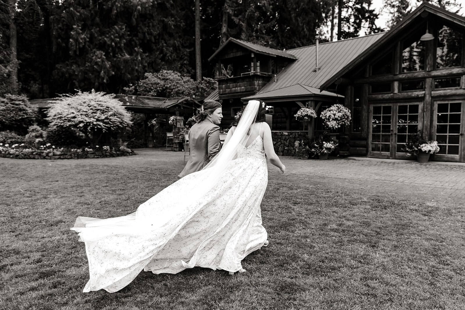 two brides walk across a lawn with dress trailing behind them