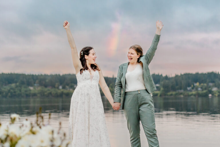 two brides cheer while standing in front of water at sunset