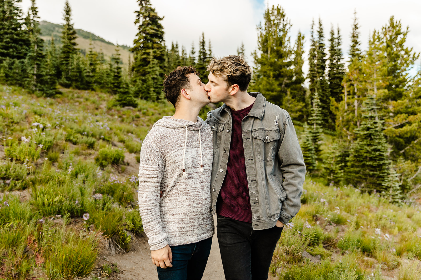 two boys kiss in a field with trees around them during mt rainier engagement session