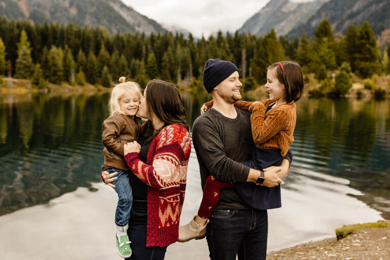 woman and man hold two little kids while standing in front of a lake