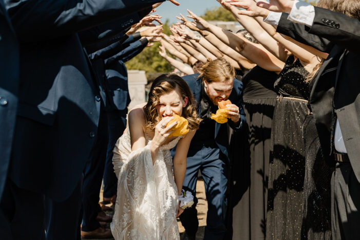 bride and groom run through tunnel of bridal party while eating sandwiches