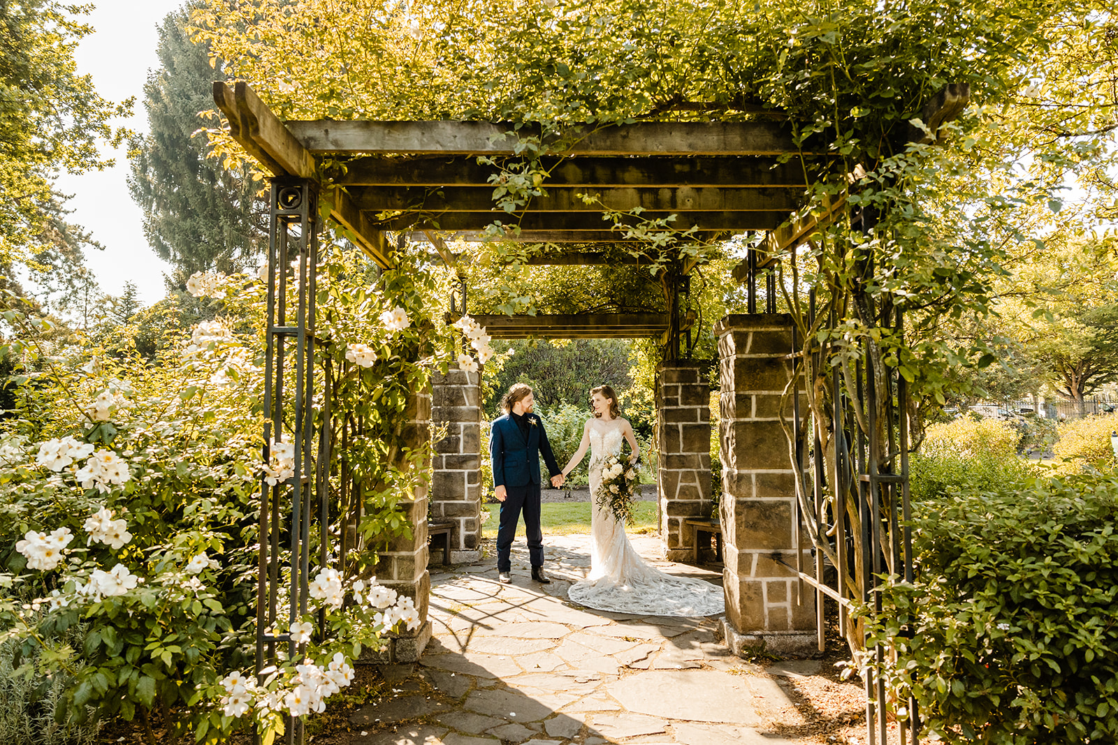 bride and groom stand under trellis in rose garden