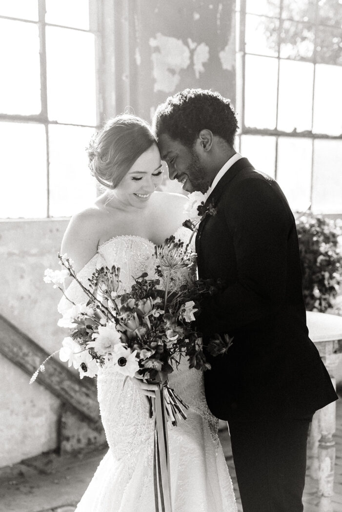 bride and groom stand forehead to forehead in front of windows