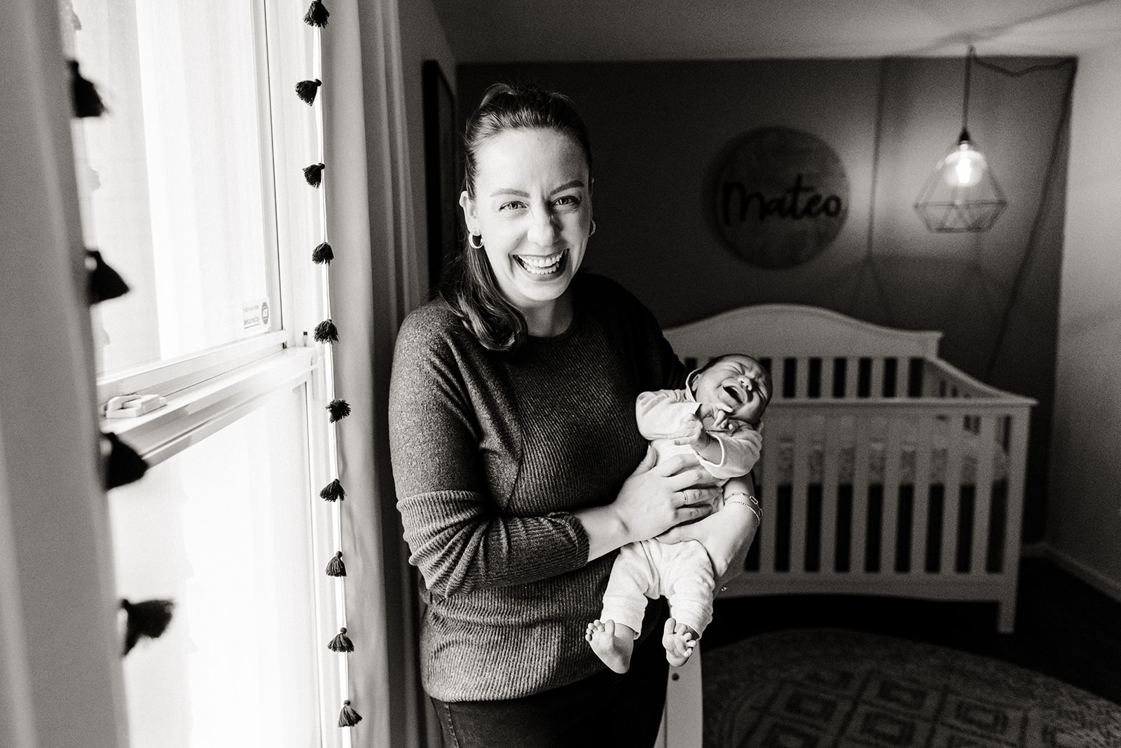 mom stands by window and laughs while baby cries in her arms
