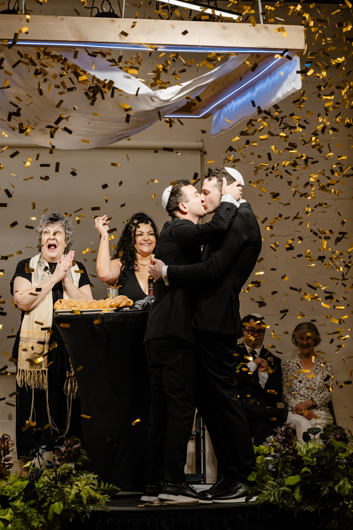 two grooms kissing under chuppah alter and gold confetti