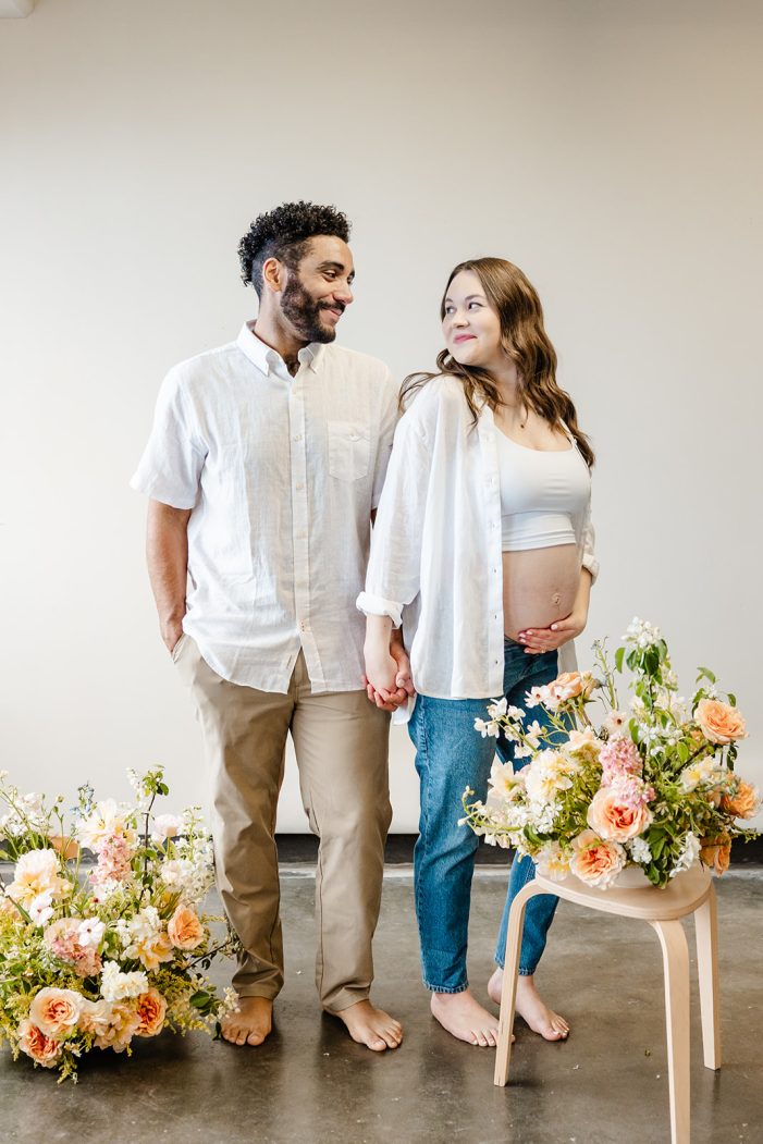 man and pregnant woman stand in studio amongst flowers